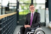 Dennis Petschner in dunklem Anzug mit lilafarbener Krawatte, im Rollstuhl sitzend an Geländer. Klick öffnet eine vergrößerte Ansicht.