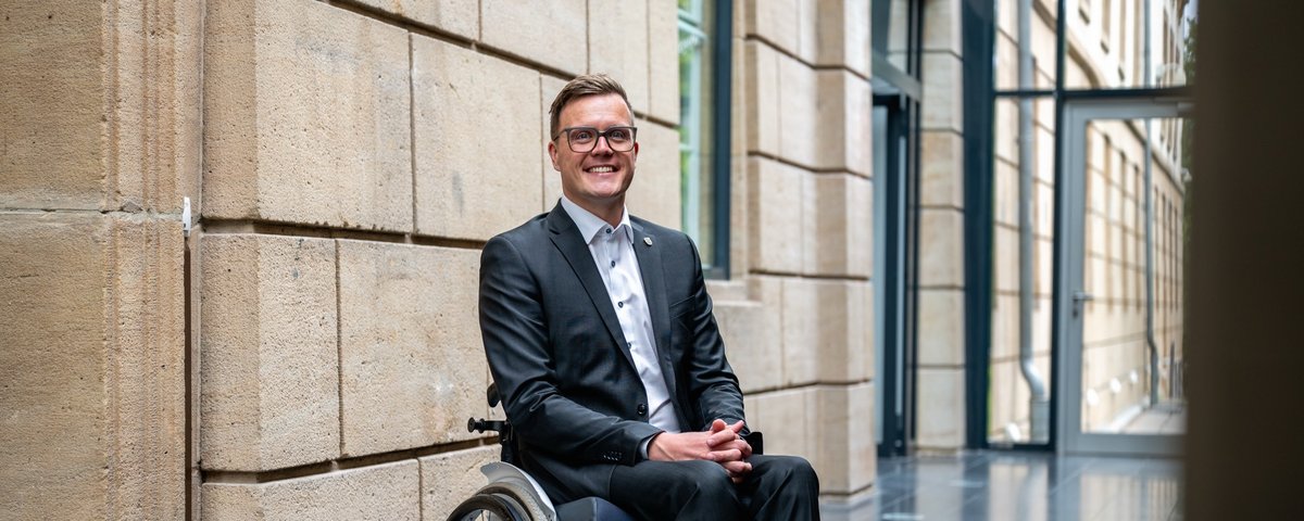 Dennis Petschner im Anzug, im Rollstuhl sitzend vor einer sandsteinfarbenen Wand im Landtag, im Hintergrund verglaste Fronten und Glastüren.