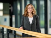 Porträtbild von Carolin Urban mit Brille und langen lockigen Haaren in grauem Anzug mit weißem T-Shirt am Geländer stehend. Klick öffnet eine vergrößerte Ansicht.