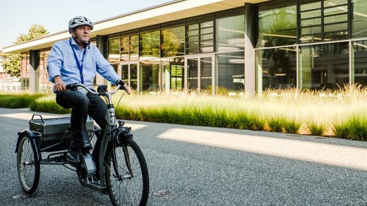 Mann auf dreirädrigem Fahrrad und mit Helm fährt an einem Büro- oder Fabrikgebäude vorbei