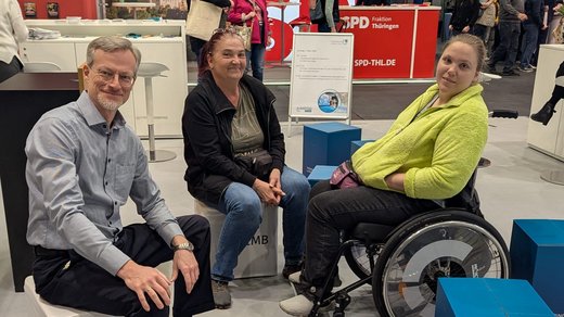 drei Personen auf blauen Hockern sitzend am Stand des TLMB: Markus Lorenz, stellvertretender TLMB, mit 2 Besucherinnen, davon eine im Rollstuhl sitzende Frau 