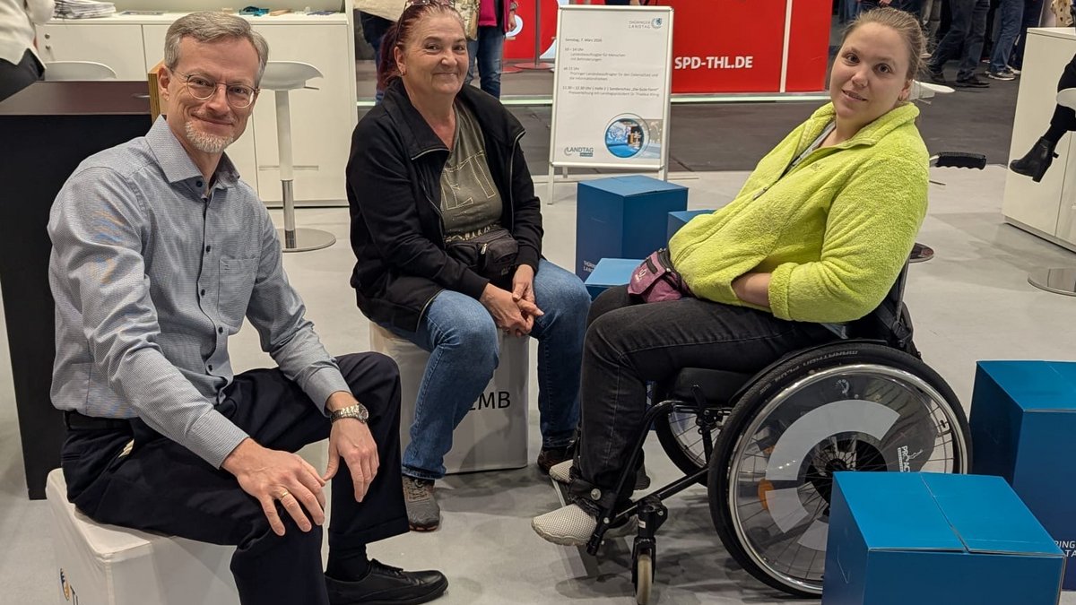Gruppe von drei Personen sitzend auf den blauen Hockern des Landtags: M. Lorenz mit zwei Besucherinnen, davon eine Frau im Rollstuhl