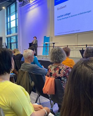 Publikumsreihen sitzend, TLMB Joachim Leibiger hält Grußwort auf der Bühne . Klick öffnet eine vergrößerte Ansicht.