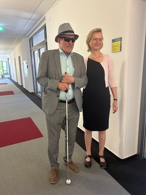 TLMB Joachim Leibiger mit Hut, Sonnenbrille und Langstock und Ministerin Wolf in schwarzem Kleid auf einem Gang des Ministeriums. Klick öffnet eine vergrößerte Ansicht.