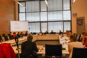 Blick in den Sitzungssaal im Landtag, Teilnehmende sitzen an langen Tischen in U-Form gestellt, Präsentationsleinwand. Blick auf Präsidium mit Moderator. Klick öffnet eine vergrößerte Ansicht.