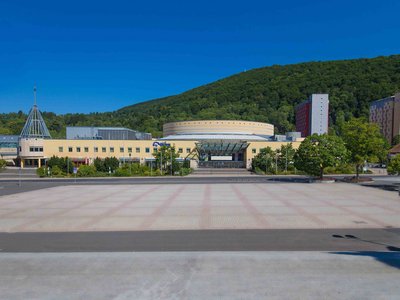 Außenansicht des CCS-Centers in Suhl, davor ein Platz und eine verlaufende Straße. Wolkenloser blauer Himmel, waldige Hügel im Hintergrund.. Klick öffnet eine vergrößerte Ansicht.