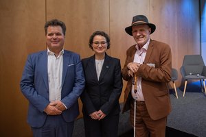 Jürgen Dusel mit AKT-Präsidentin Ines M. Jauck und TLMB Joachim Leibiger vor hölzerner Wand. Klick öffnet eine vergrößerte Ansicht.