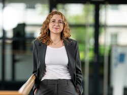 Porträtbild von Carolin Urban mit langen Haaren und Brille, im grauen Anzug, am Geländer und vor einer Glastür. Klick öffnet eine vergrößerte Ansicht.