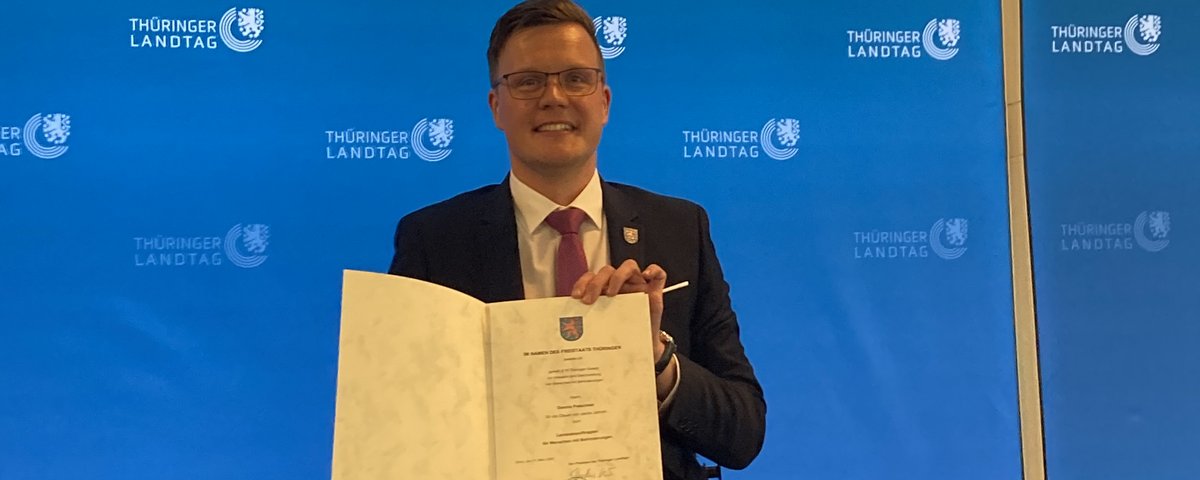 Dennis Petschner mit Ernennungsurkunde vor blauer Fotowand mit dem Logo des Thüringer Landtags