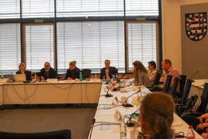 Blick in den Sitzungssaal im Landtag, Teilnehmende sitzen an langen Tischen in U-Form. Blick auf Präsidium mit Moderator. Klick öffnet eine vergrößerte Ansicht.