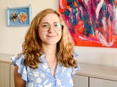 Porträtbild von Carolin Urban mit langen Haaren und Brille , in hellblauer Bluse vor einem abstrakten Gemälde im Besprechungsraum des TLMB. Klick öffnet eine vergrößerte Ansicht.