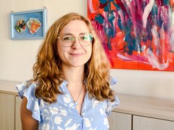 Porträtbild von Carolin Urban mit langen Haaren und Brille , in hellblauer Bluse vor einem abstrakten Gemälde im Besprechungsraum des TLMB. Klick öffnet eine vergrößerte Ansicht.