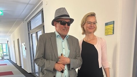 Joachim Leibiger mit Hut und Sonnebrille neben Ministerin Katja Wolf in schwarzem Kleid, beide stehen auf einem Gang des Ministeriums