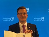 Dennis Petschner mit Brille und in dunklem Anzug die Ernennungsurkunde in den Händen, aufgenommen vor der blauen Fotowand des Thüringer Landtags. Klick öffnet eine vergrößerte Ansicht.