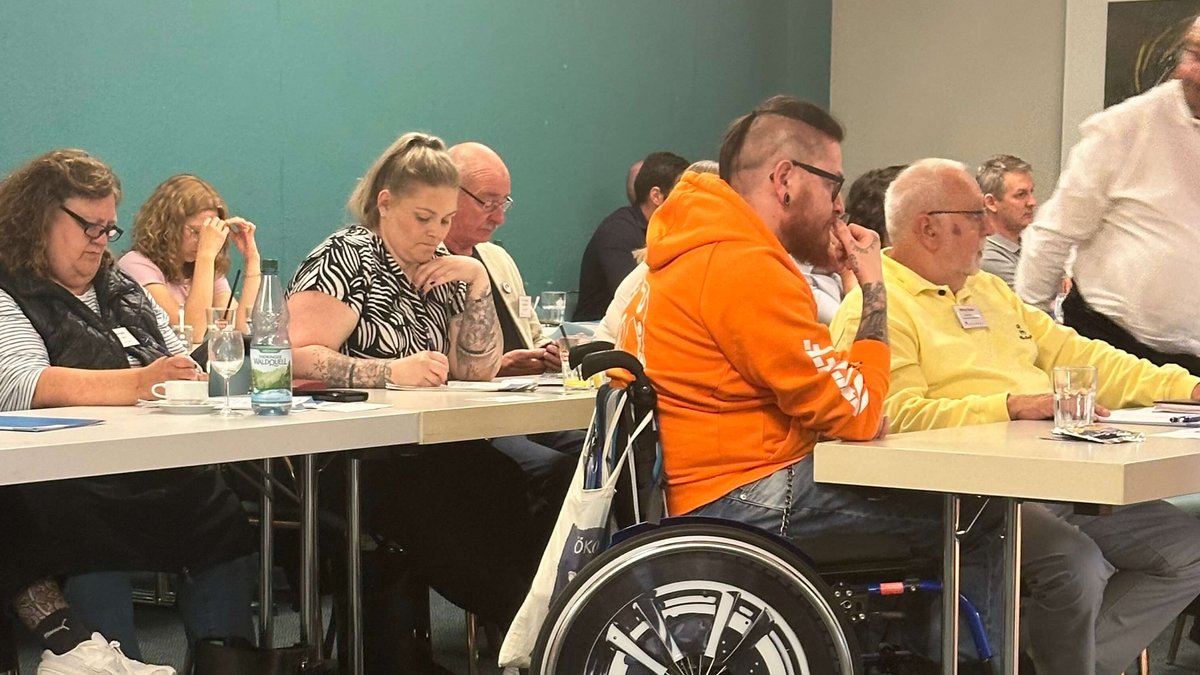 Teilnehmende an Tischen, Mann mit Brille, im orangen Pulli und im Rollstuhl am Tisch 